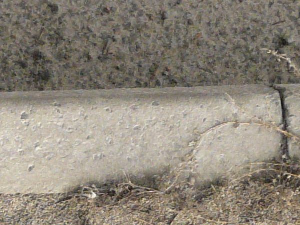 Road edge texture, with grey and white asphalt separated from a cracked sidewalk by a pattern left by melted snow. Brown weeds and fine dust cover the surface of the sidewalk.